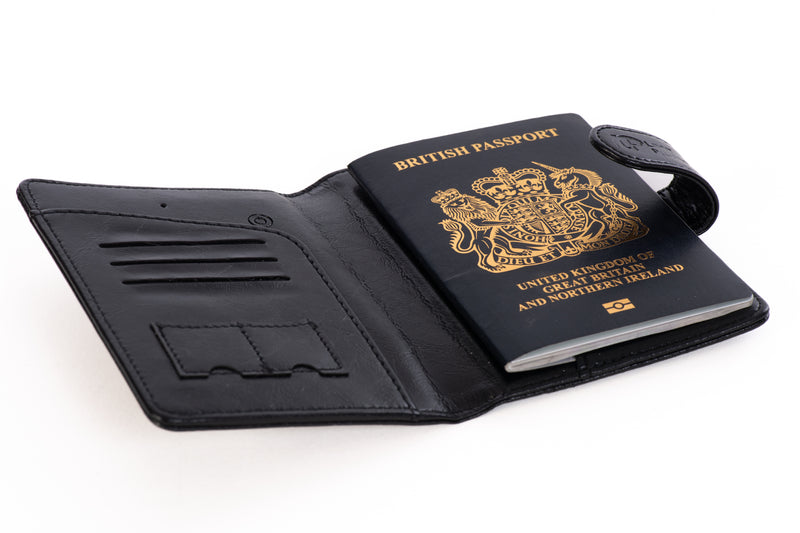 "Angled view of the LostProof CardPass, a black leather GPS-tracked passport holder. The holder is open, displaying a British passport securely placed in the right-side pocket. The left side features multiple card slots and additional compartments for organizing travel essentials. The snap-button closure strap, embossed with the LostProof logo, is open, highlighting the secure yet accessible design.
