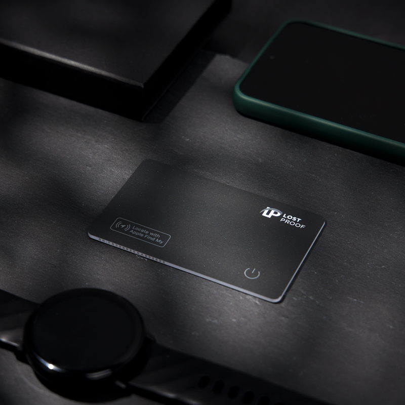 LostProof CardTrack wallet tracker placed on a dark granite surface, surrounded by a smartwatch, smartphone, and a black box. A sleek, minimalistic design with Apple Find My compatibility.