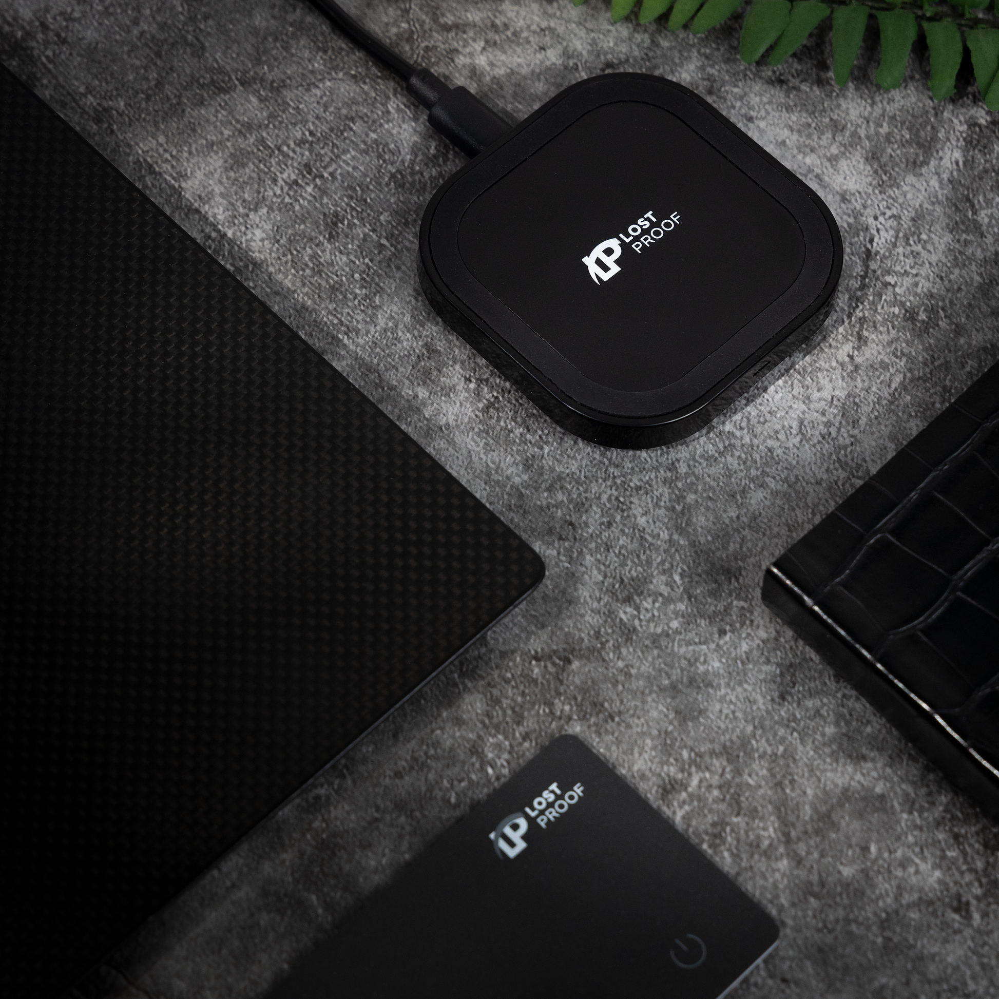 Black electronic wireless charger device with a brand logo 'LostProof' on a grey surface with a plant in the background.