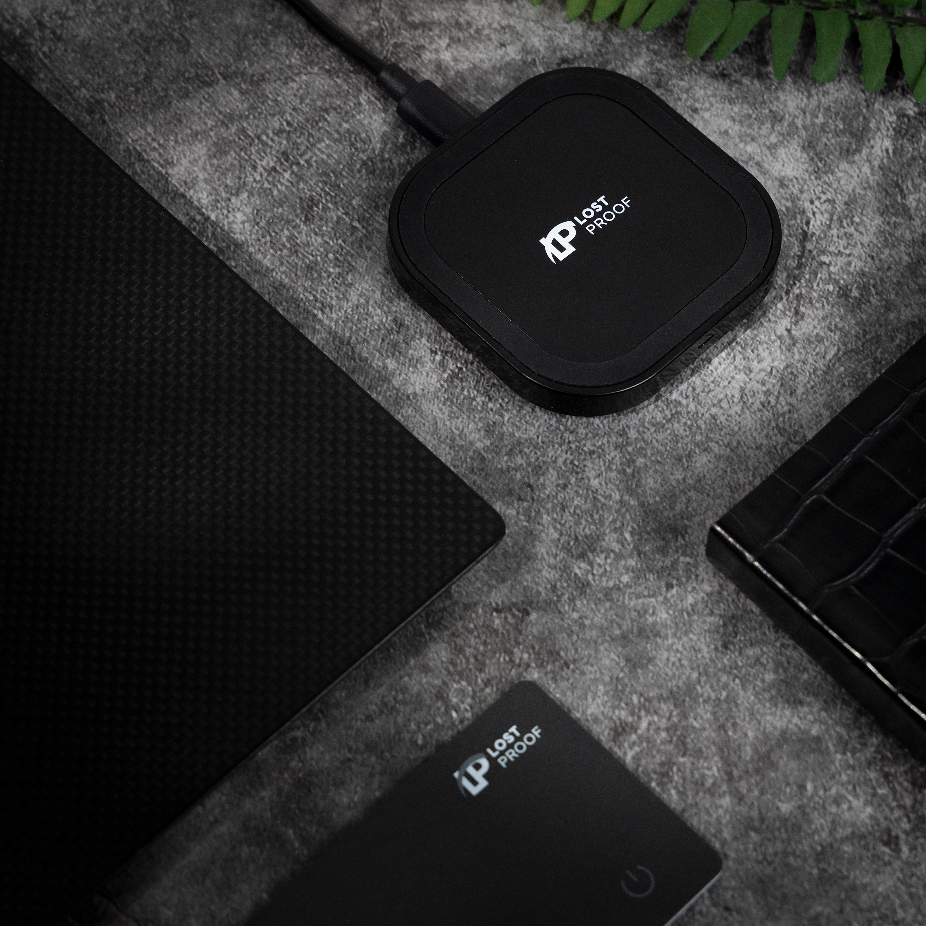 Black electronic wireless charger device with a brand logo 'LostProof' on a grey surface with a plant in the background.
