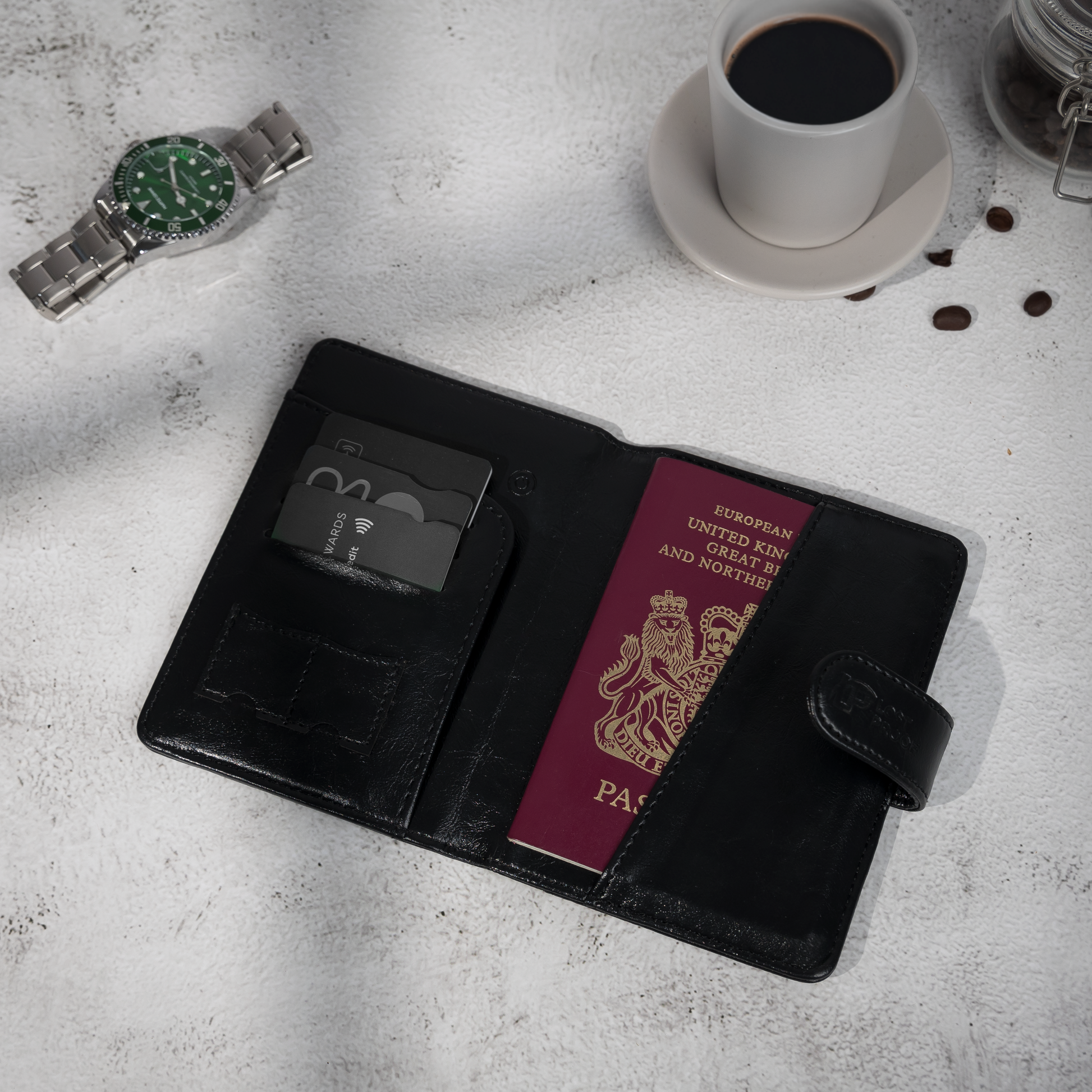 Stylish flat-lay of the LostProof CardPass, a black leather GPS-tracked passport holder, placed on a textured surface. The holder is open, revealing a British passport partially tucked into the right-side pocket and multiple bank cards neatly organized in the left-side card slots. A stainless steel wristwatch and a cup of black coffee with scattered coffee beans are arranged beside it, adding a sophisticated travel aesthetic.
