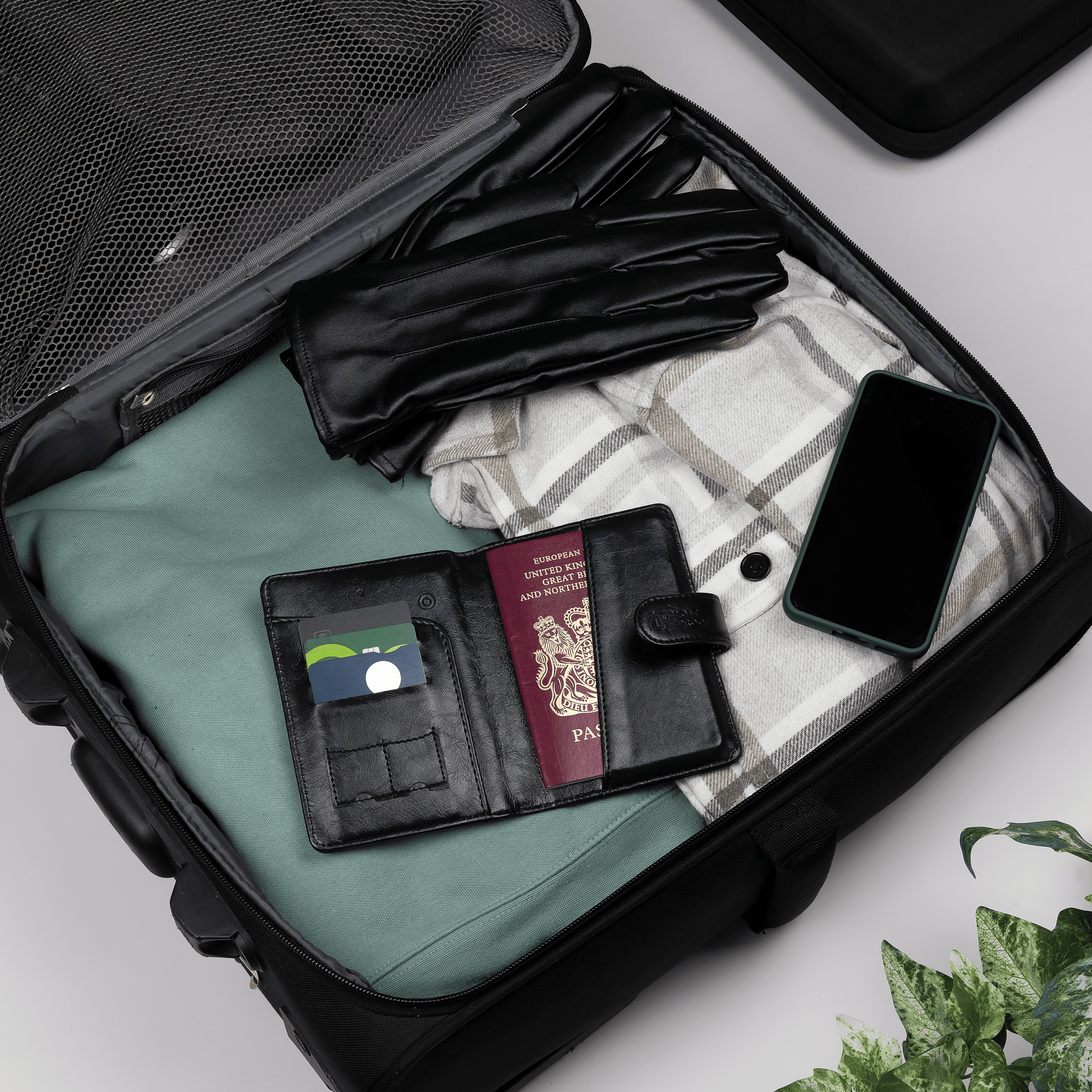 LostProof black GPS-tracked passport holder open inside a packed suitcase, holding a UK passport and cards, alongside travel essentials like gloves, a phone, and a blanket.