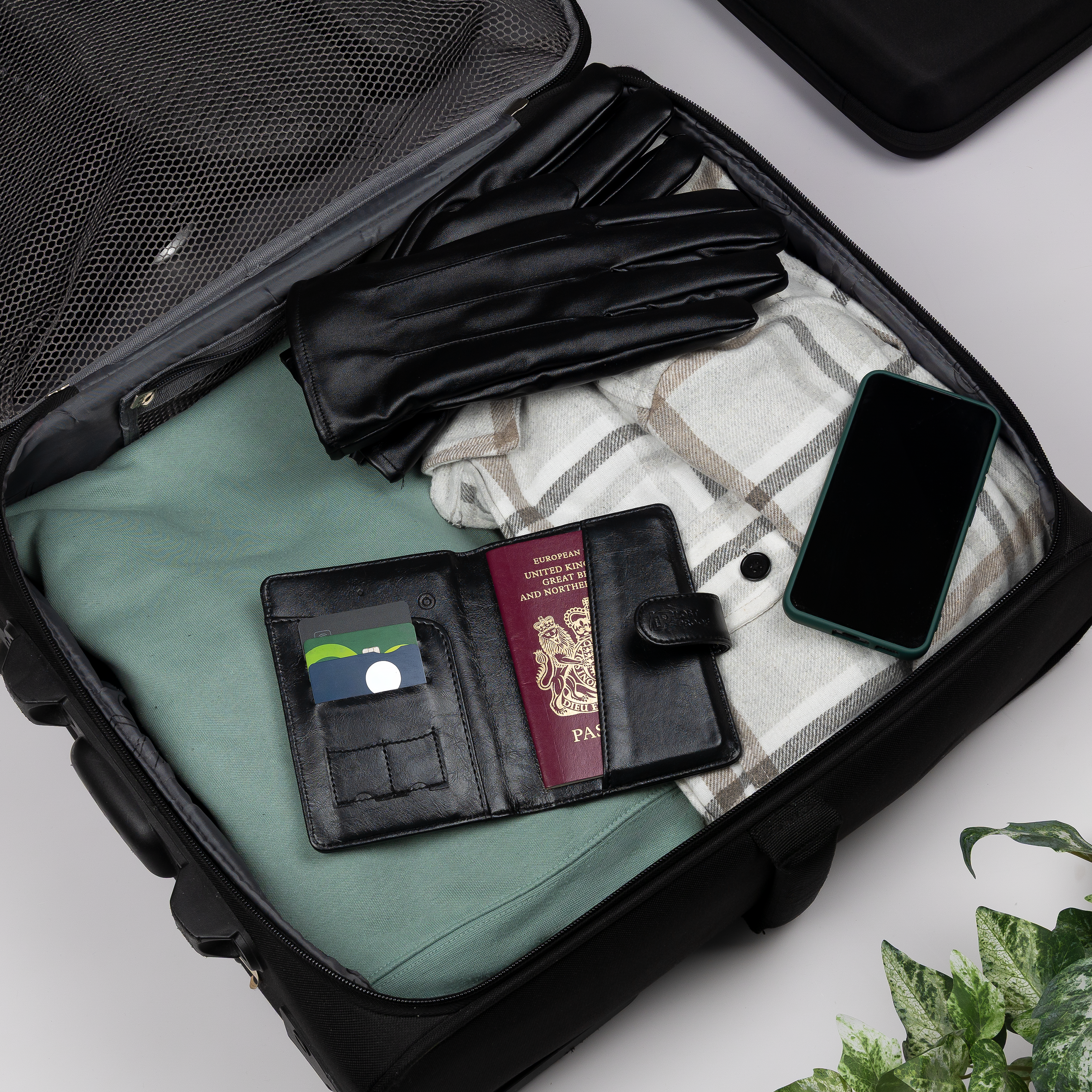 LostProof black GPS-tracked passport holder open inside a packed suitcase, holding a UK passport and cards, alongside travel essentials like gloves, a phone, and a blanket.